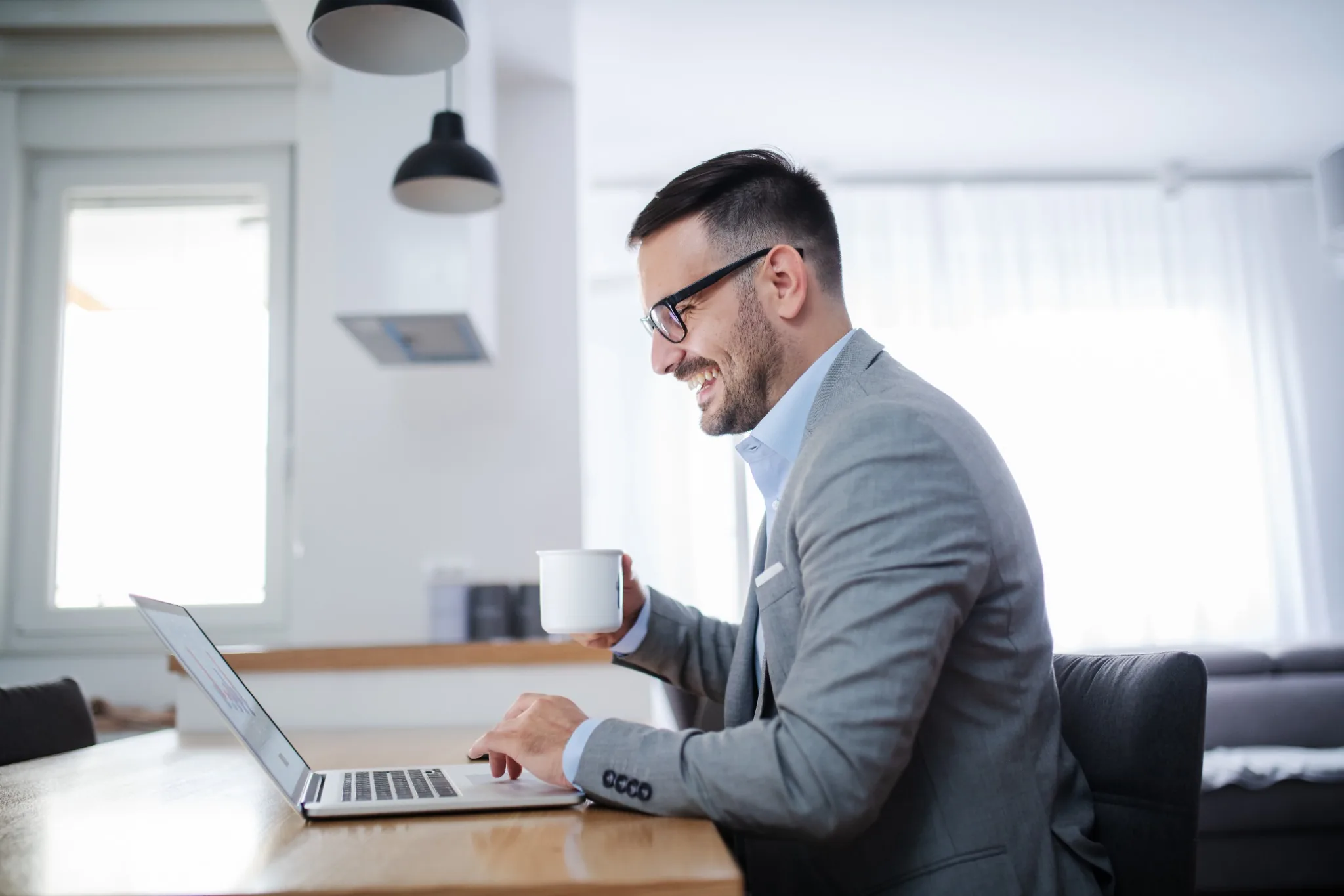 Happy man in suit sitting at a desk holding a cup of coffee and looking at his laptop